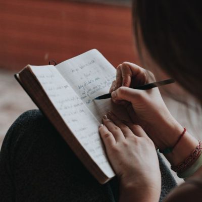 Sitzende Frau mit einem geöffneten Heft, hält ein Stift in der Hand und schreibt